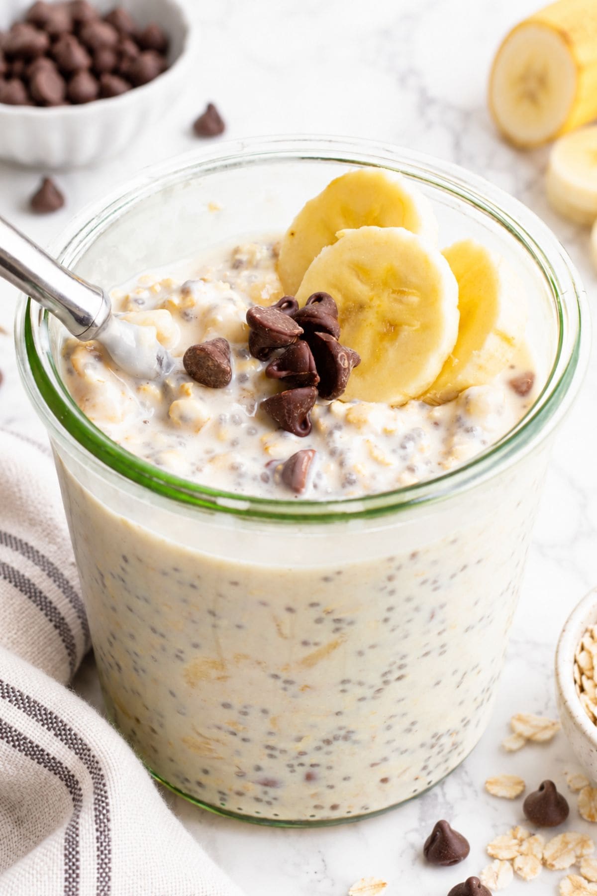 Glass jar of banana chocolate chip overnight oats with chia seeds, topped with banana slices and chocolate chips, a spoon resting in the jar, and a striped linen towel beside it.