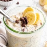 Glass jar of banana chocolate chip overnight oats with chia seeds, topped with banana slices and chocolate chips, a spoon resting in the jar, and a striped linen towel beside it.