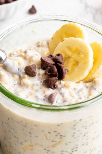 Glass jar of banana chocolate chip overnight oats with chia seeds, topped with banana slices and chocolate chips, a spoon resting in the jar, and a striped linen towel beside it.