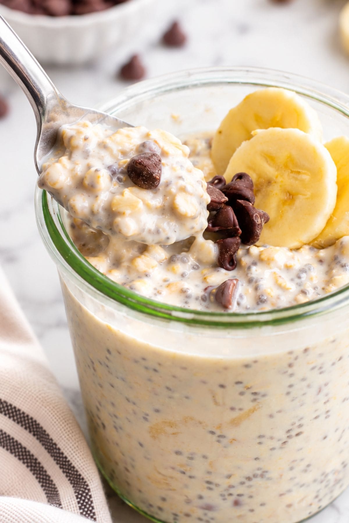 Spoonful of chewy oats, chia seeds, and a chocolate chip lifted from a jar of overnight oats garnished with banana slices.