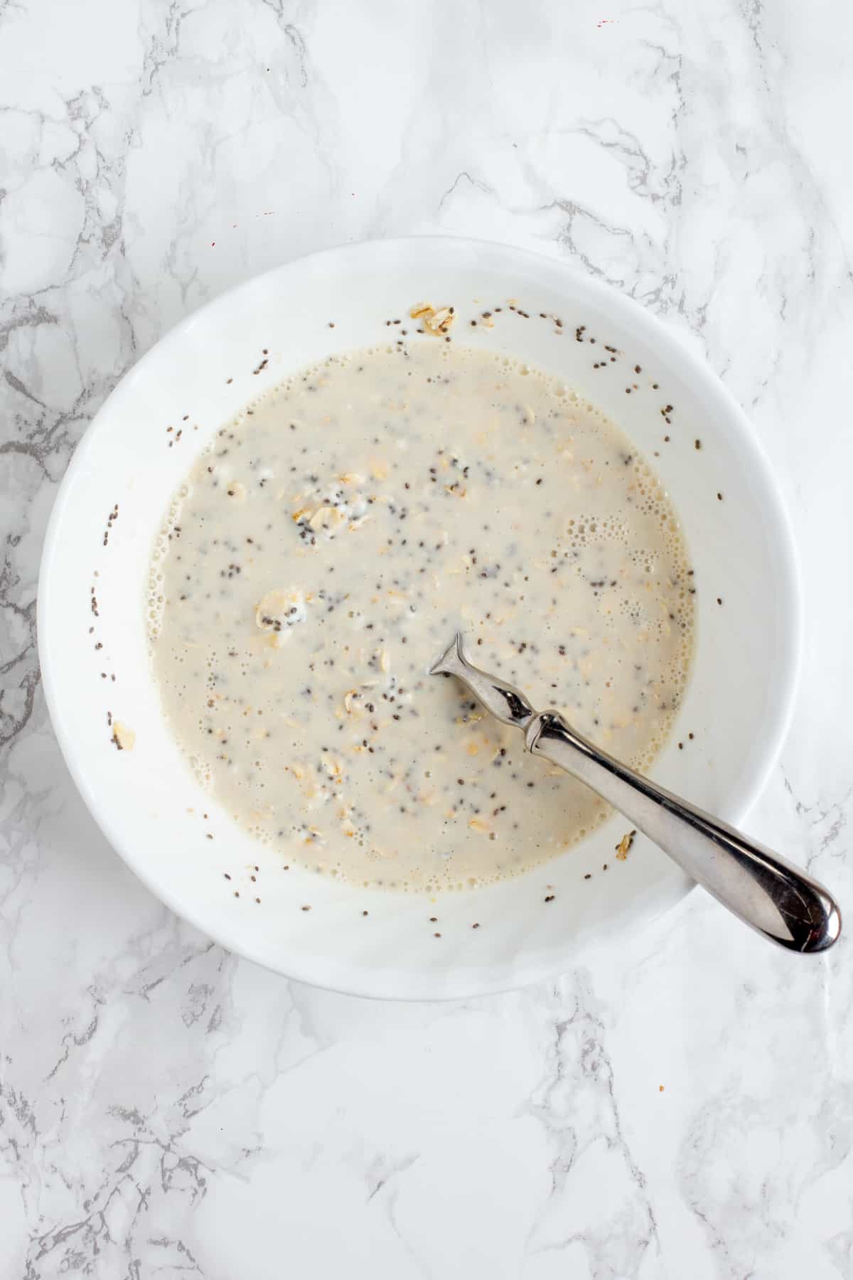 Stirring together the ingredients for overnight oats in a small white mixing bowl with a spoon. The mixing bowl in on a gray marble countertop.