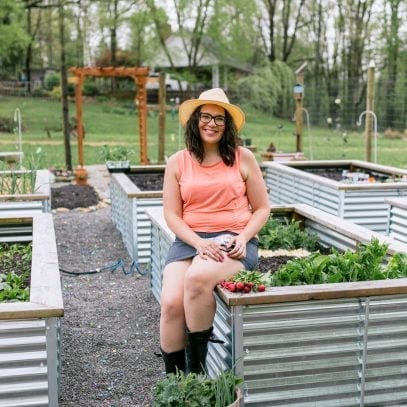 Brunette woman in a coral shirt and a sun hat sitting on the edge of a raised garden bed