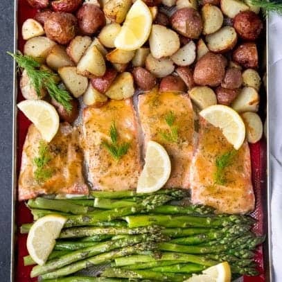 Overhead of a sheet pan with a silicon baking sheet covered with roasted salmon, asparagus, potatoes, fresh lemon slices, and fresh herbs.
