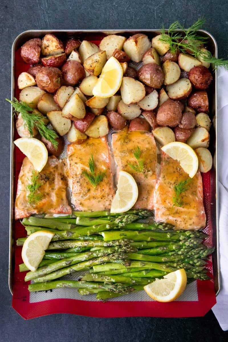 Overhead of a sheet pan with a silicon baking sheet covered with roasted salmon, asparagus, potatoes, fresh lemon slices, and fresh herbs.