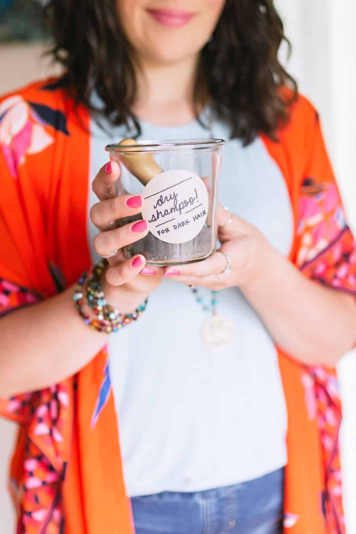 Brunette woman in a neutral top and orange kimono holding a jar labeled "dry shampoo" in both hands.