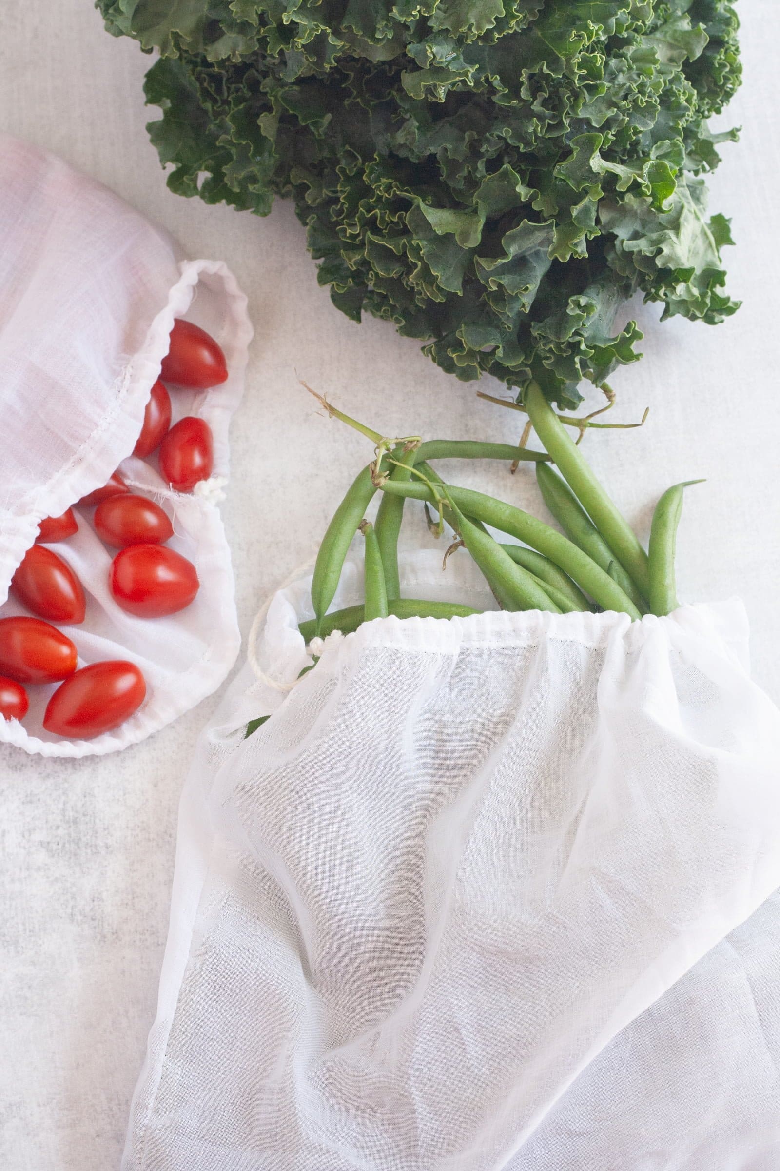 Three white cotton voile reusable produce bags filled with kale, cherry tomatoes, and green beans.