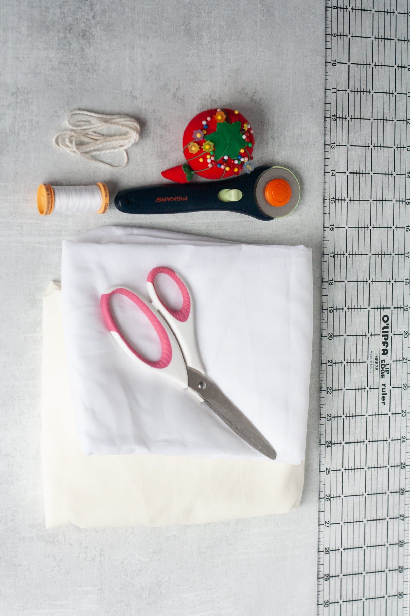 Materials for reusable produce bags laid out on a cutting mat - cotton voile, scissors, a rotary cutter, pincushion, drawstring, thread, and ruler