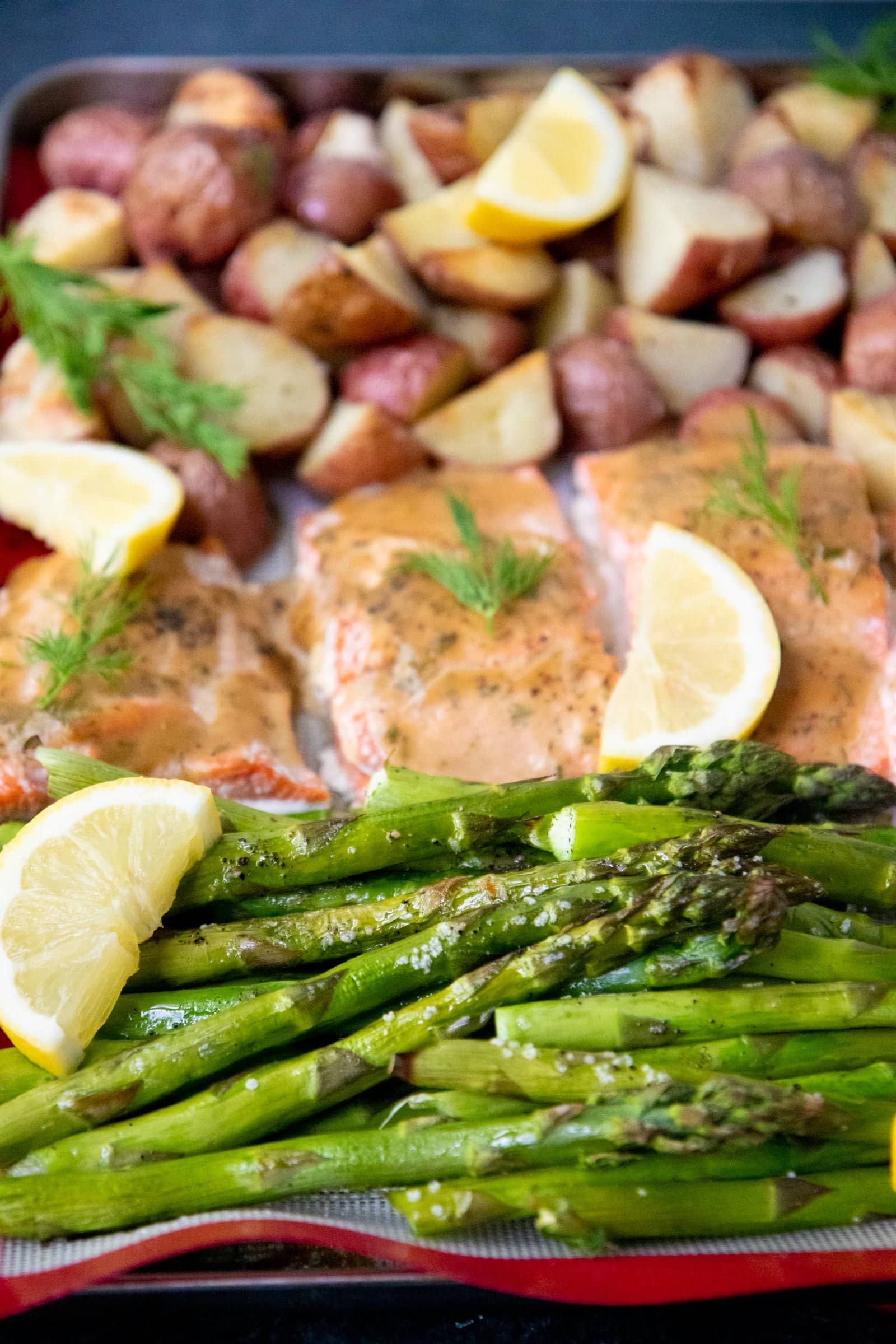 Close-up of roasted asparagus, salmon, and potatoes arranged on a baking sheet with fresh lemon and herbs.