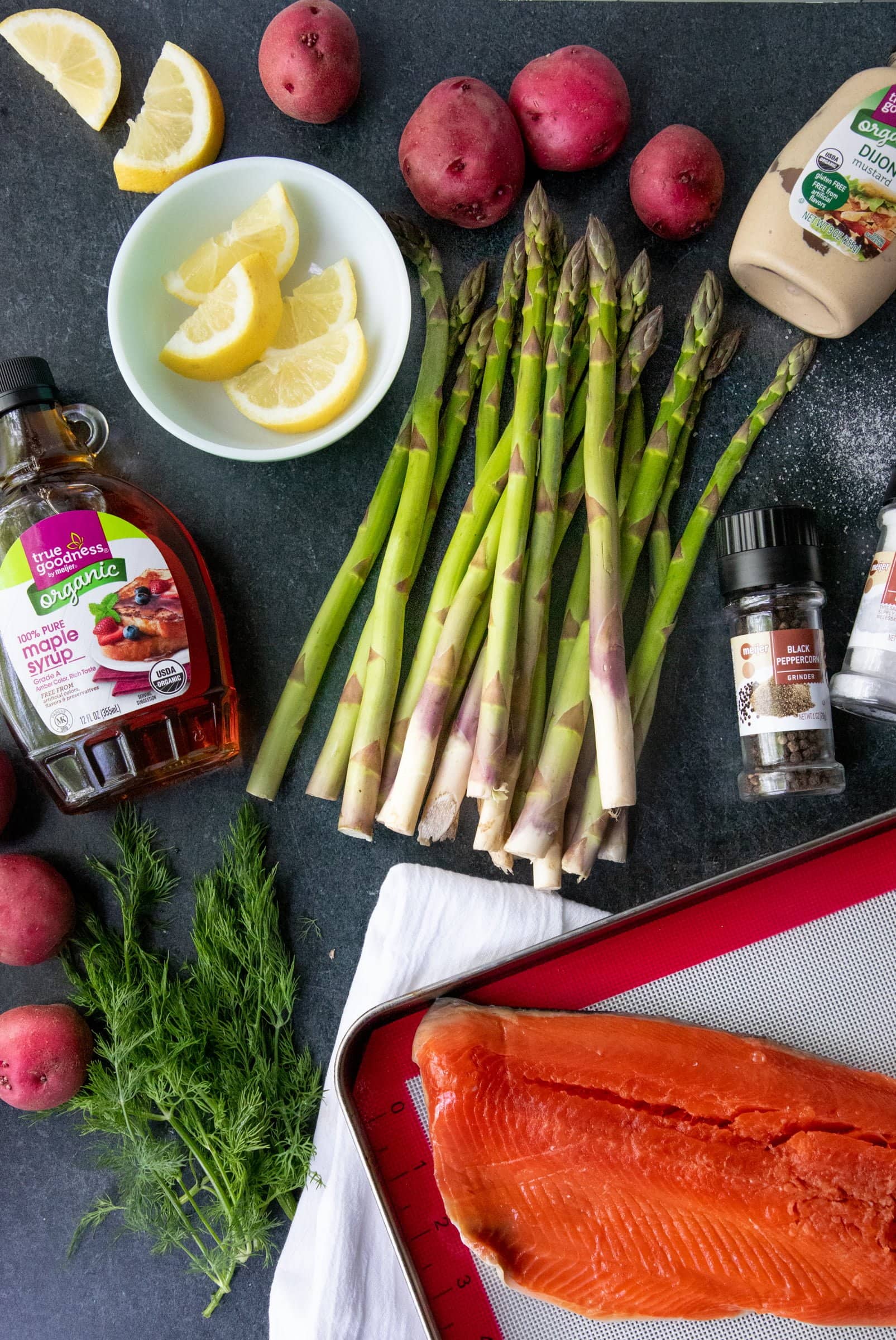 Overhead of ingredients needed, including salmon fillet, asparagus spears, red potatoes, maple syrup, dijon mustard, lemon slices, fresh dill, salt, and pepper.