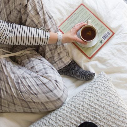 Woman in plaid pajama pants sitting on a bed with a book and mug of tea.