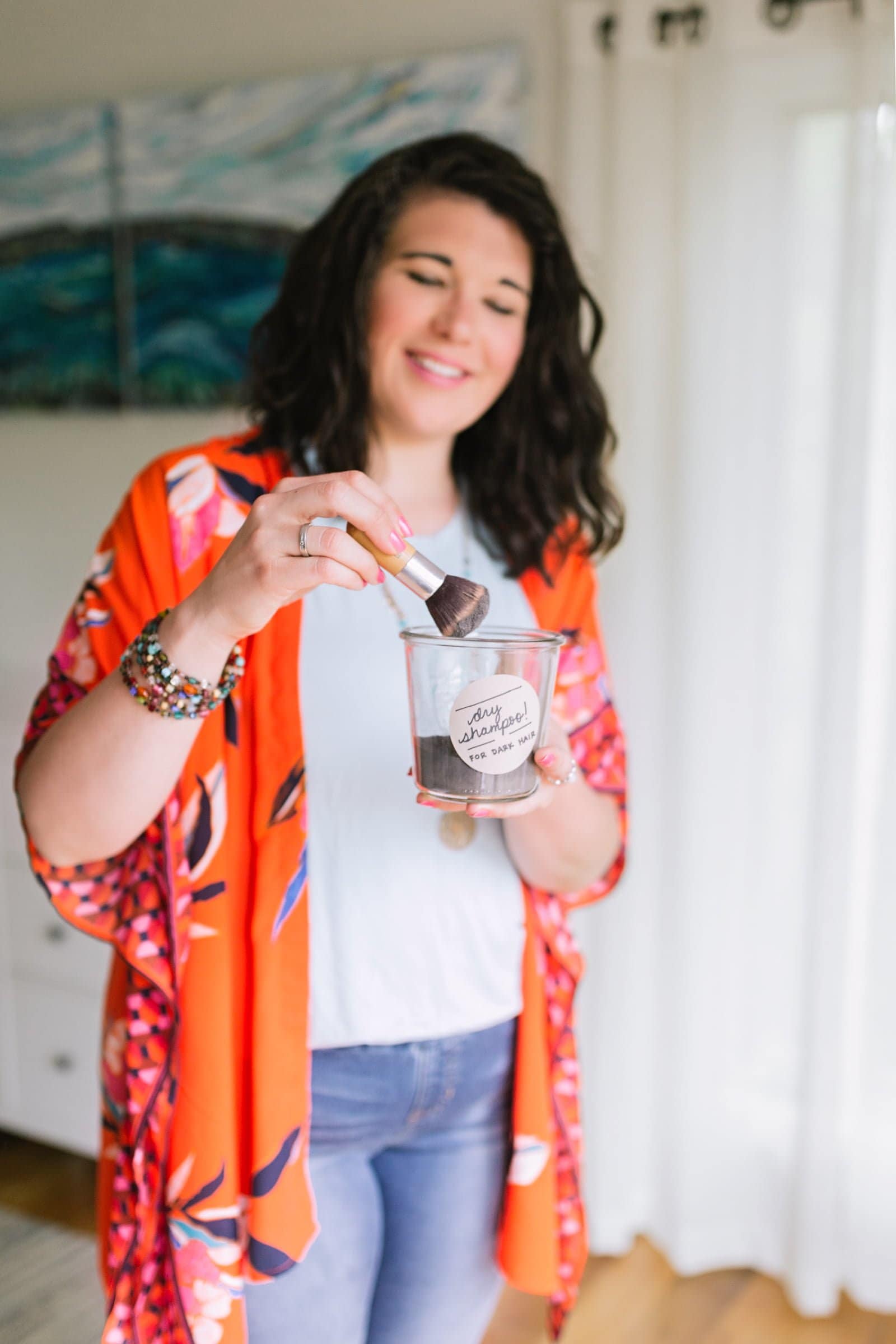 Brunette woman in a neutral top and orange kimono using a brush to mix up some DIY dry shampoo.