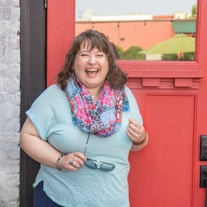Woman with curly hair in a blue top and a scarf, laughing in front of a red door.