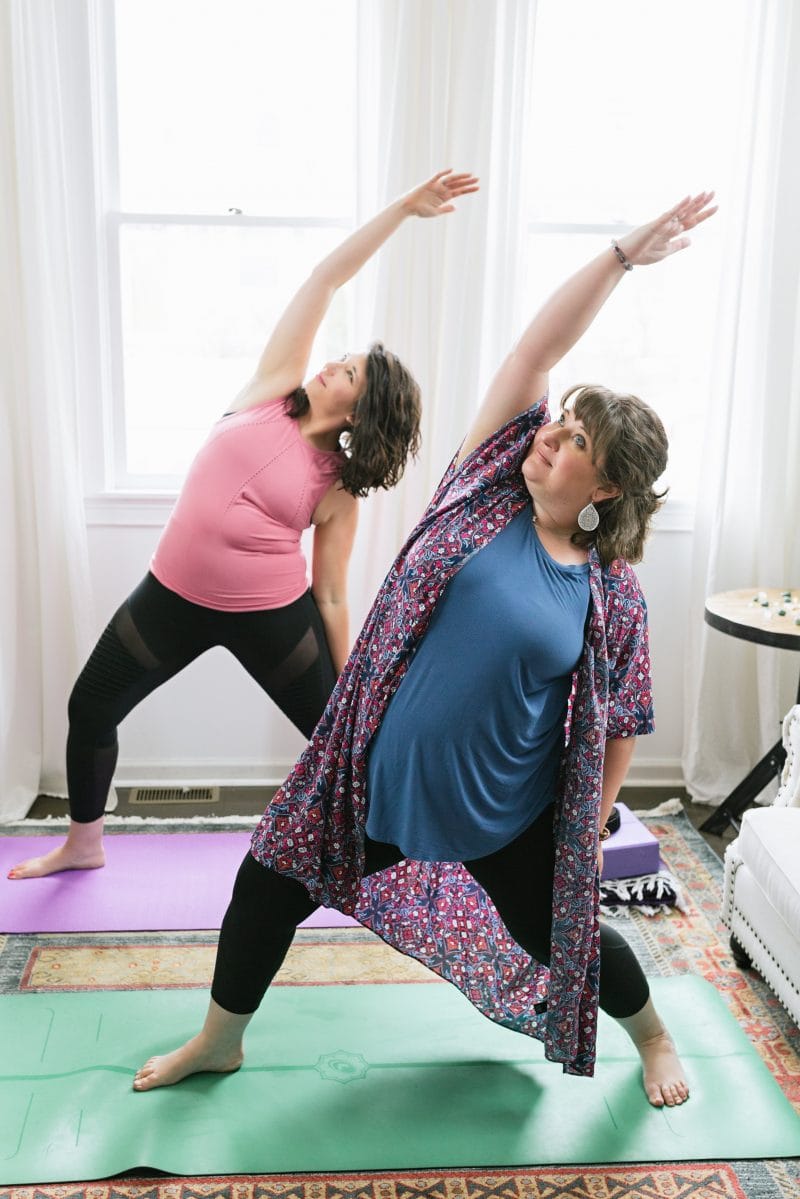 Two brunette women standing on yoga mats in a Reverse Warrior position, practicing some gentle yoga.