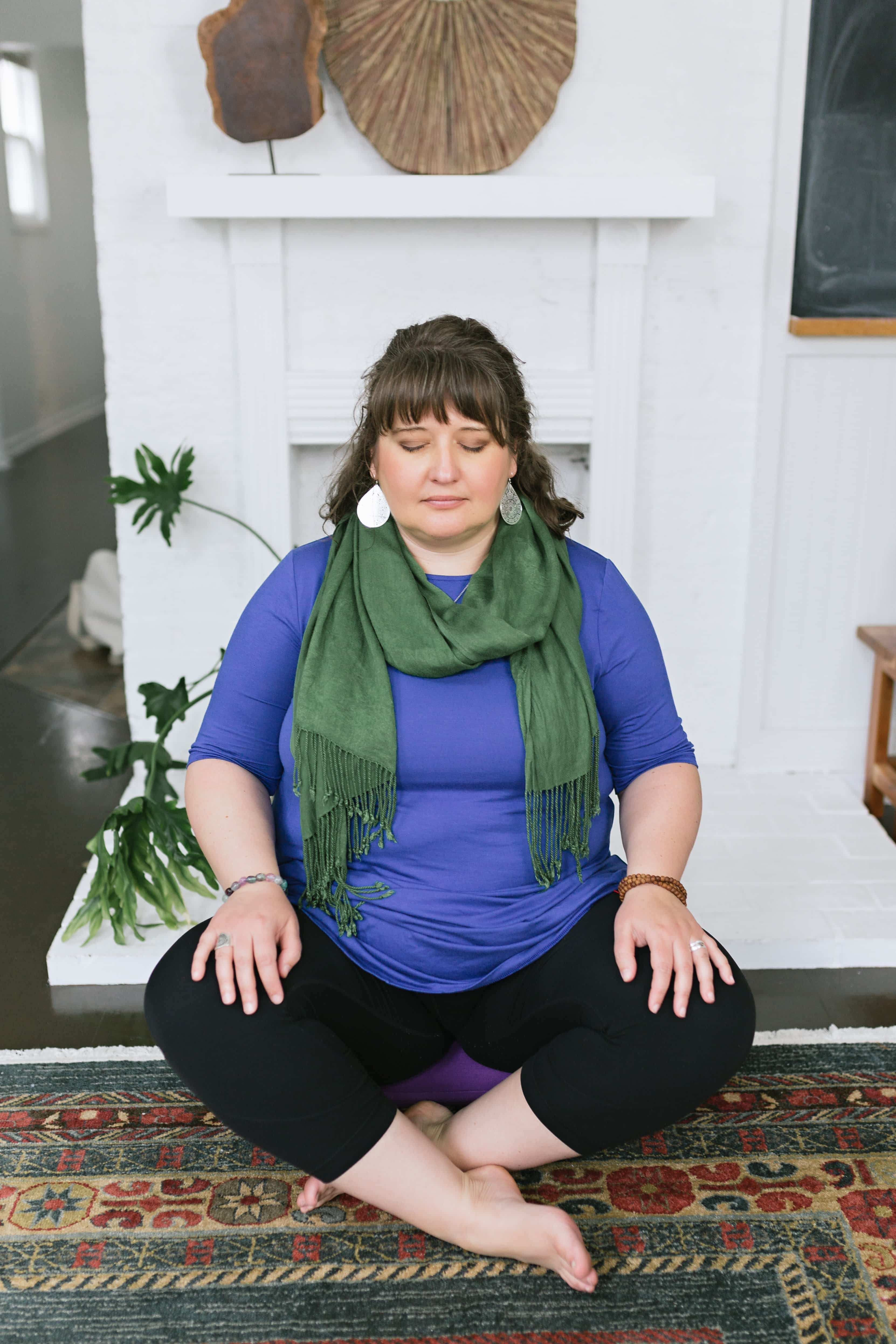 Brunette woman in a purple top and green scarf sitting cross legged with her eyes closed