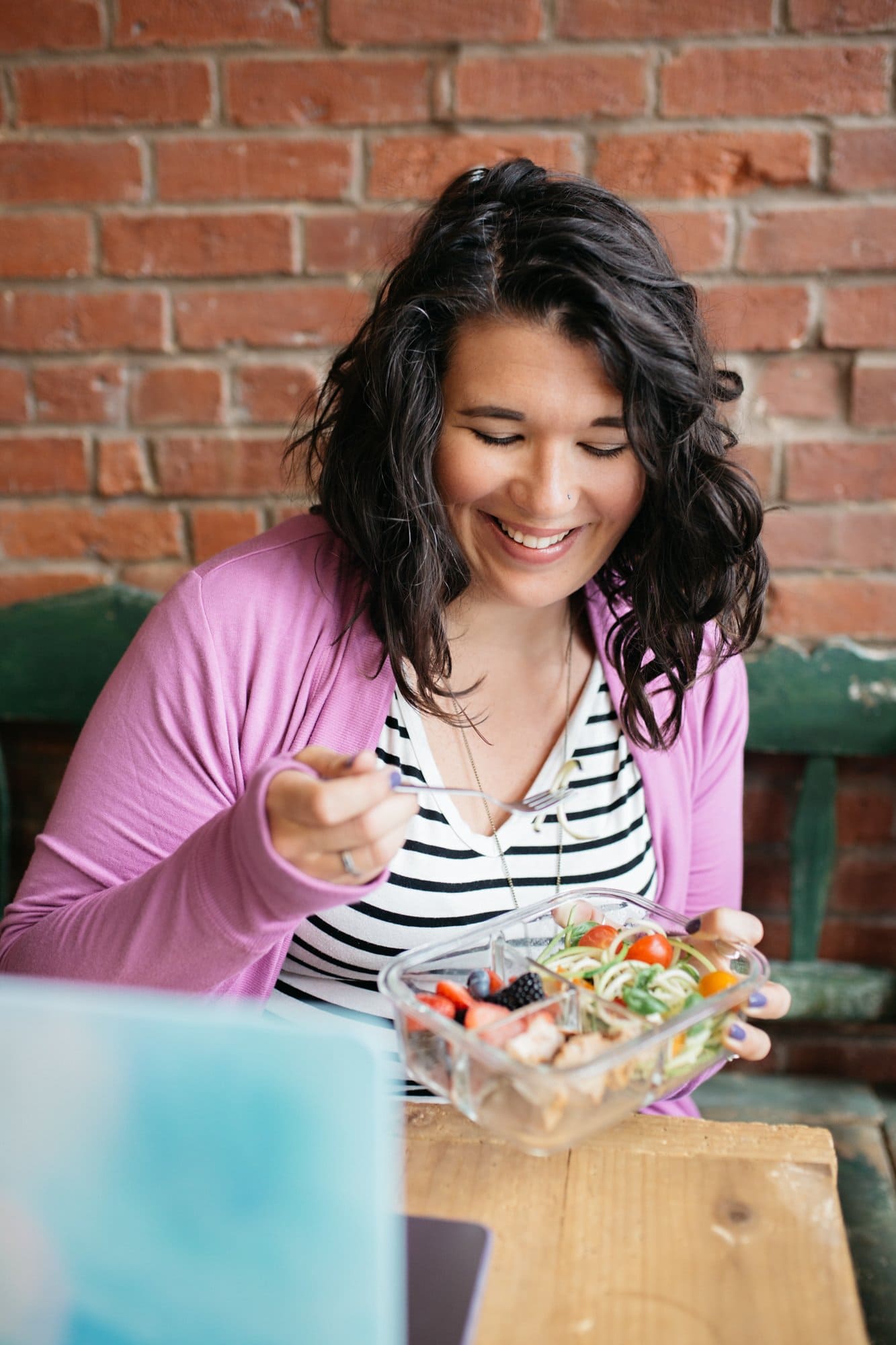 Woman in a striped shirt and purple cardigan smiling and eating vegetables, chicken, and berries out of a glass meal prep container.