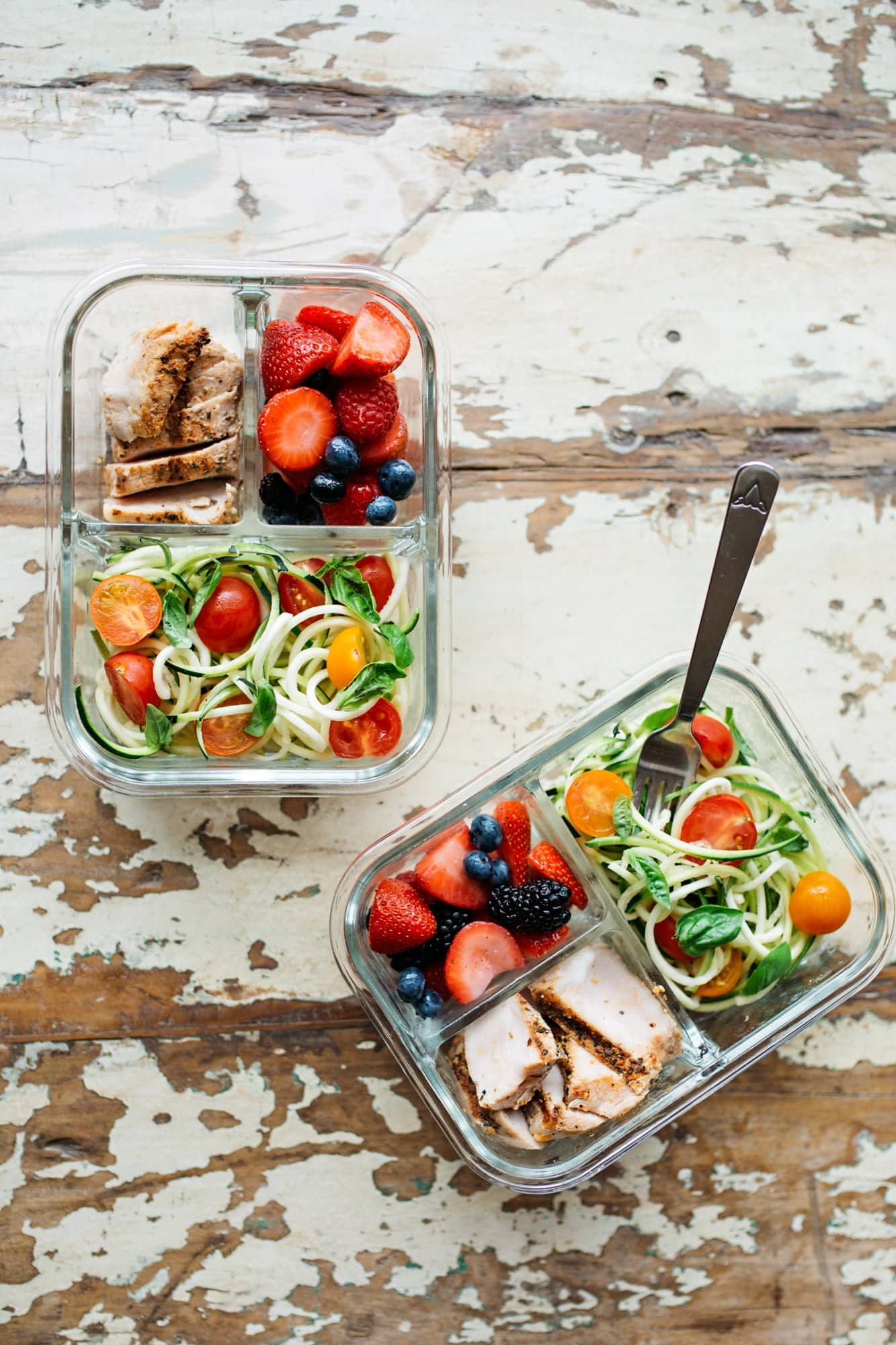Two divided glass containers filled with a meal prep lunch - chicken, berries, and vegetables.