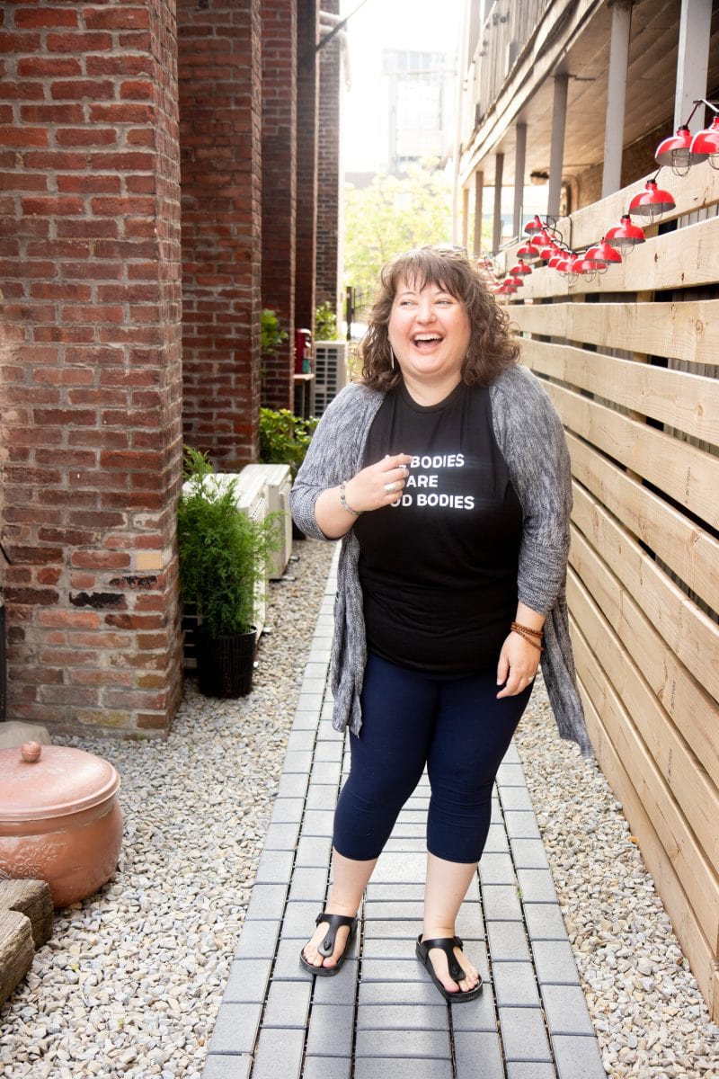 Brunette curly-haired woman standing and laughing in a shirt that says "All Bodies Are Good Bodies."