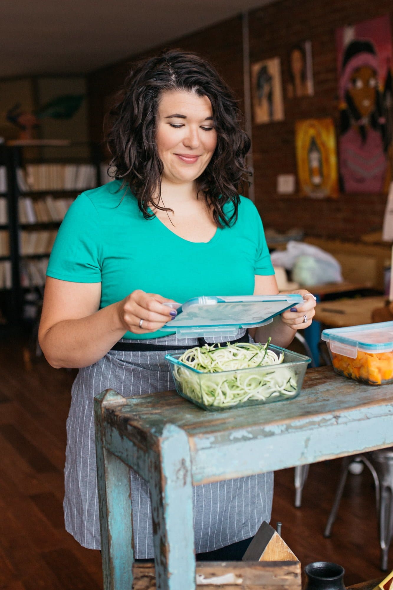 Brunette woman in a teal shirt putting the lid on a glass container filled with zucchini noodles.