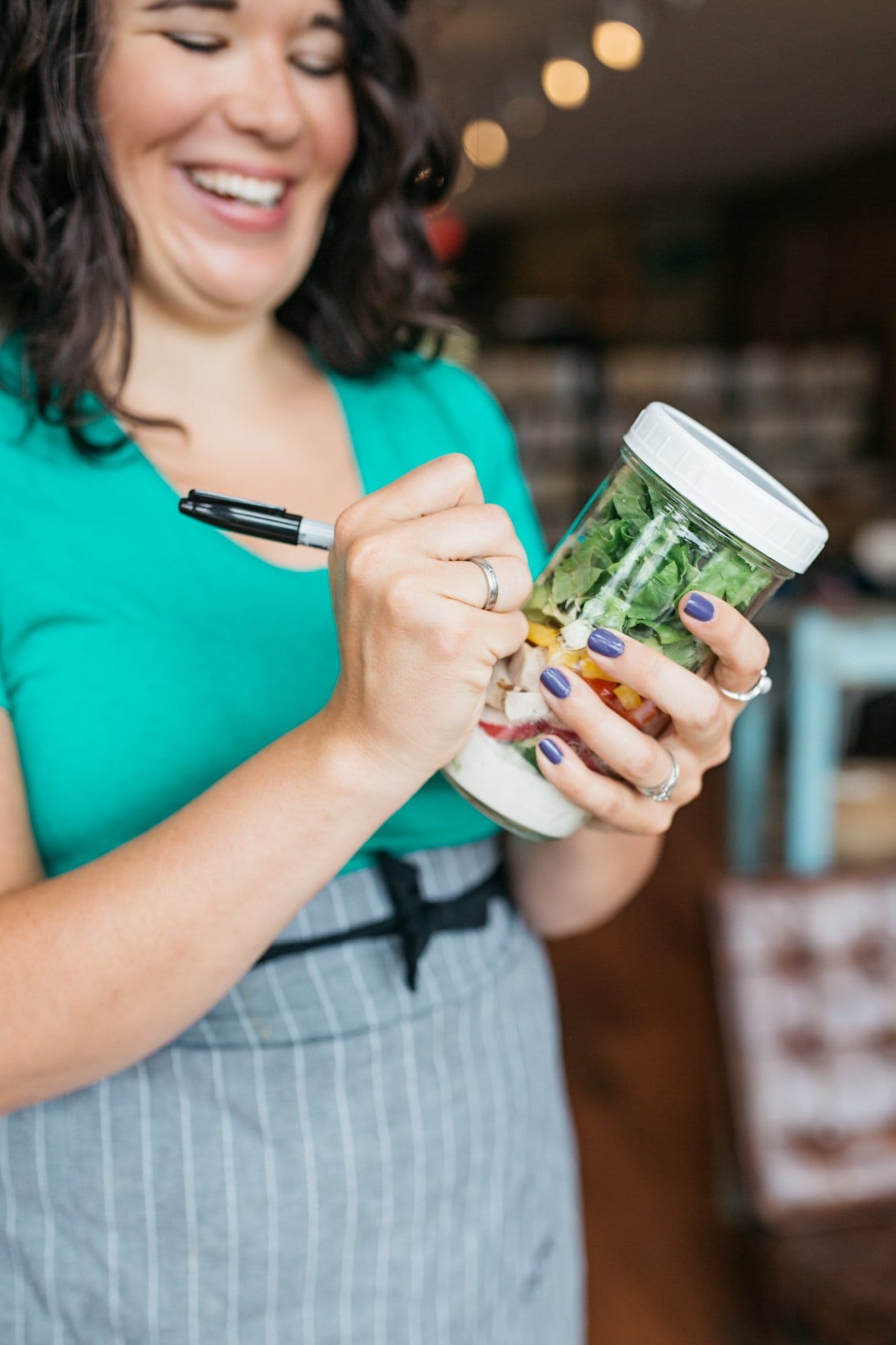 Woman in a teal shirt using a Sharpie to label a glass jar filled with salad in a jar.