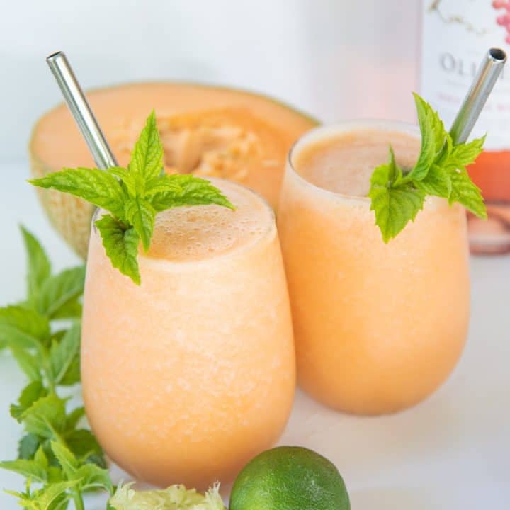 Two glasses filled with cantaloupe frosé, garnished with mint and straws. A halved cantaloupe and mint sprigs surround the glasses.