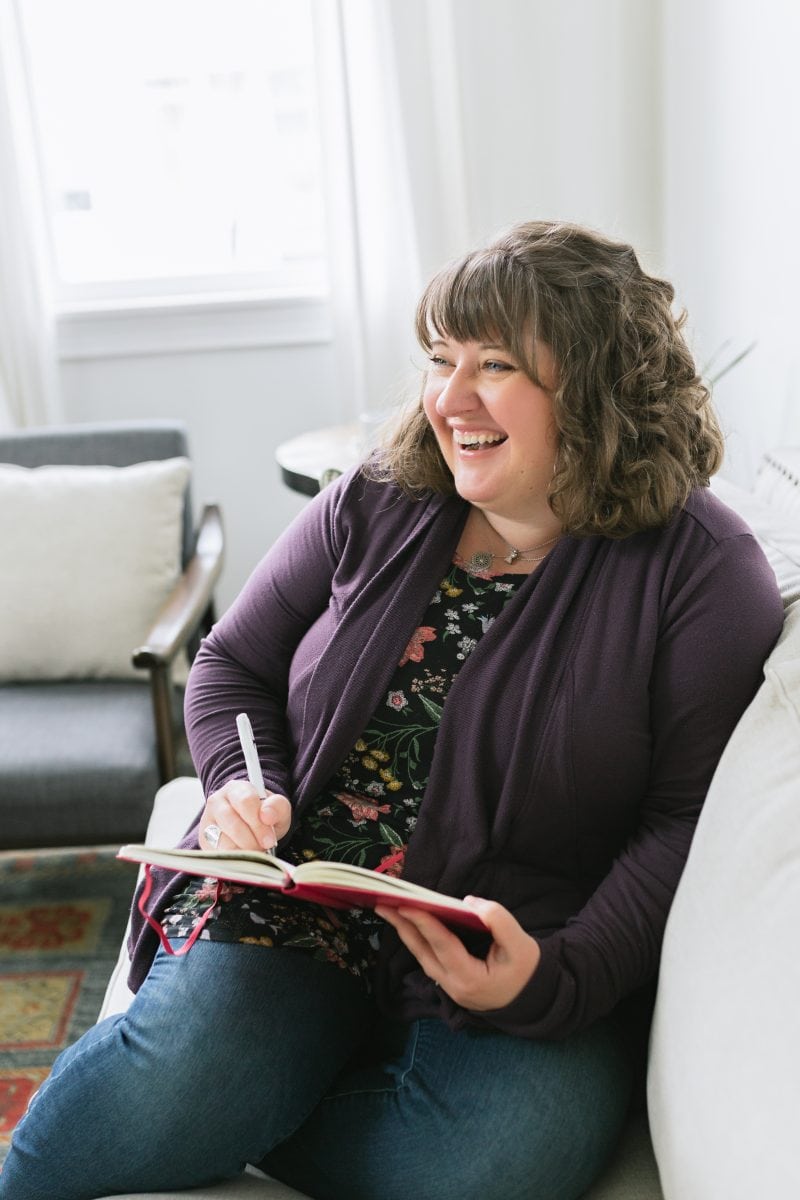 Brunette woman in a purple cardigan sitting on a white couch, laughing while she writes in a journal