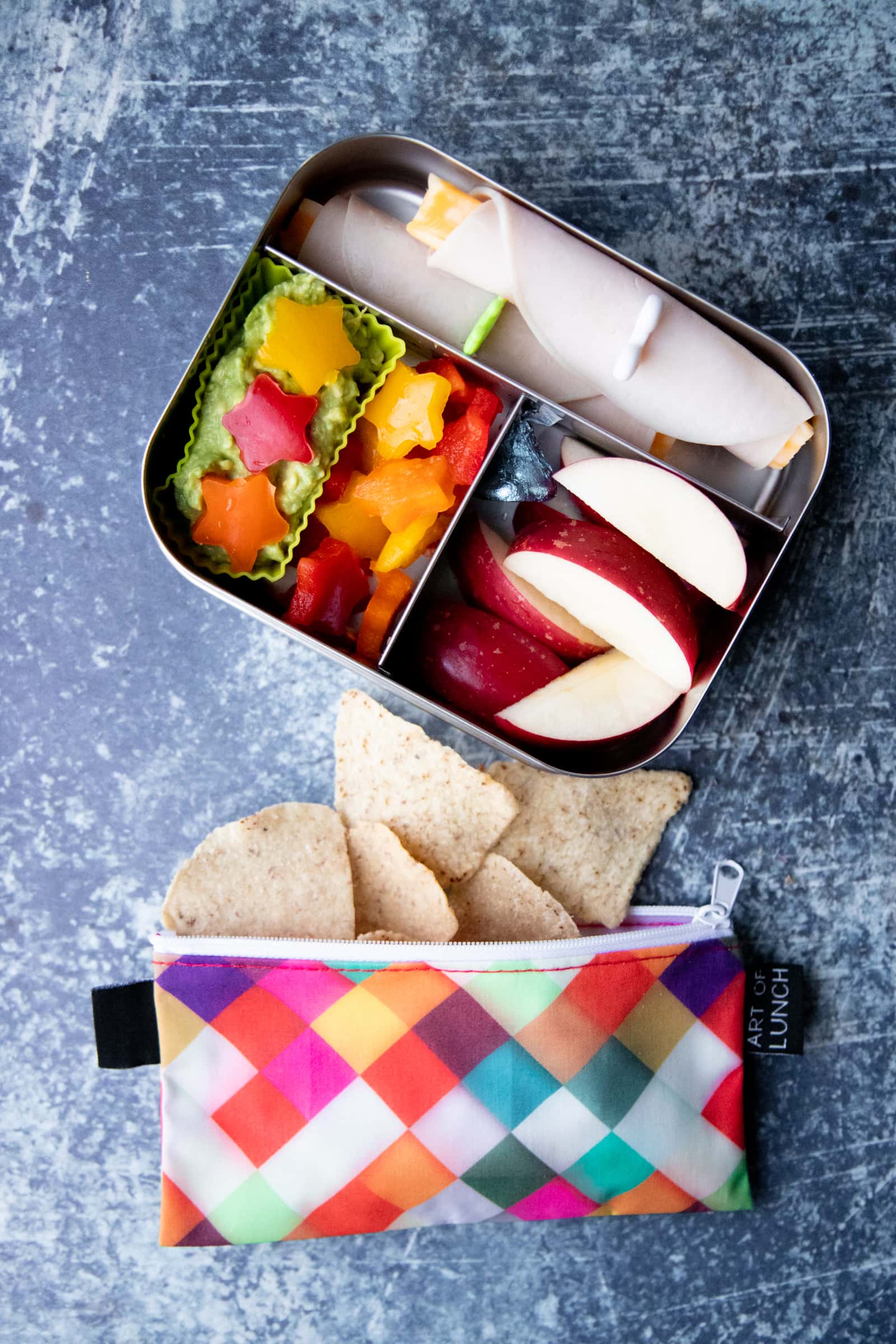Packed lunch of apple slices, bell pepper stars, guacamole, turkey and cheese roll-ups, and a bag of tortilla chips.