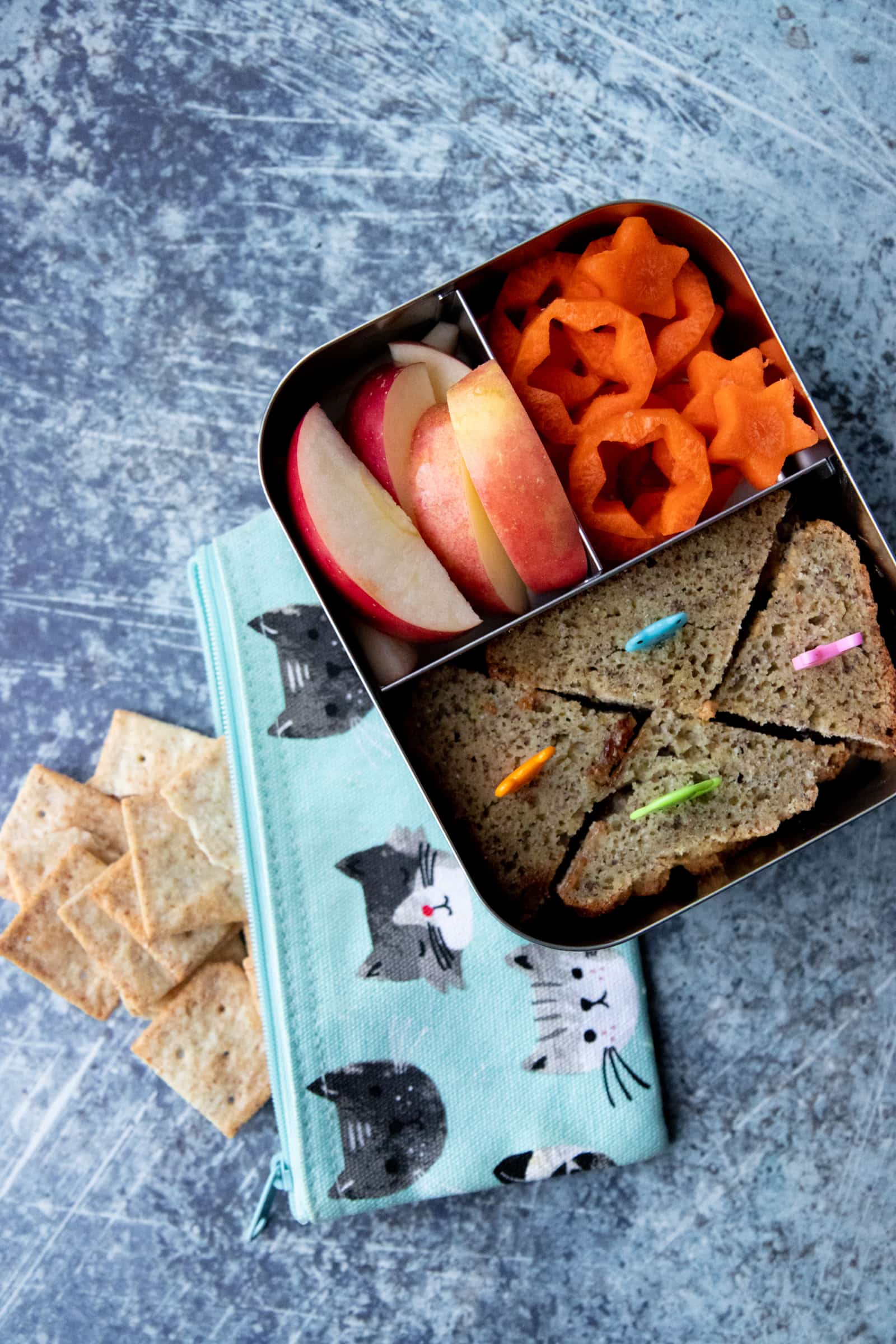 Steel lunchbox on top of a bag of crackers. The lunch box holds a sandwich, apple slices, and carrots cut into stars.