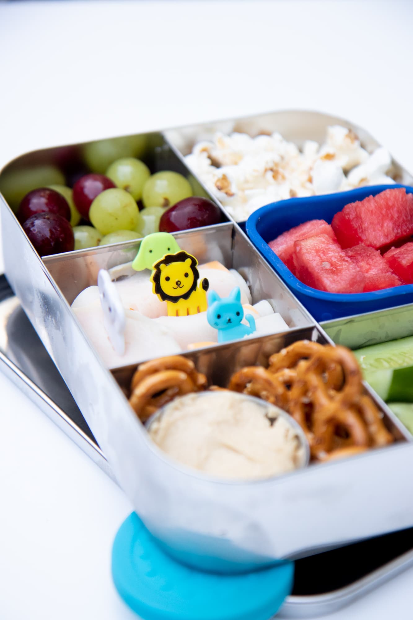 Stainless steel bento-style lunchbox filled with a snack lunch: pretzels and dip, grapes, watermelon, popcorn, cucumber spears, and deli meat roll-ups