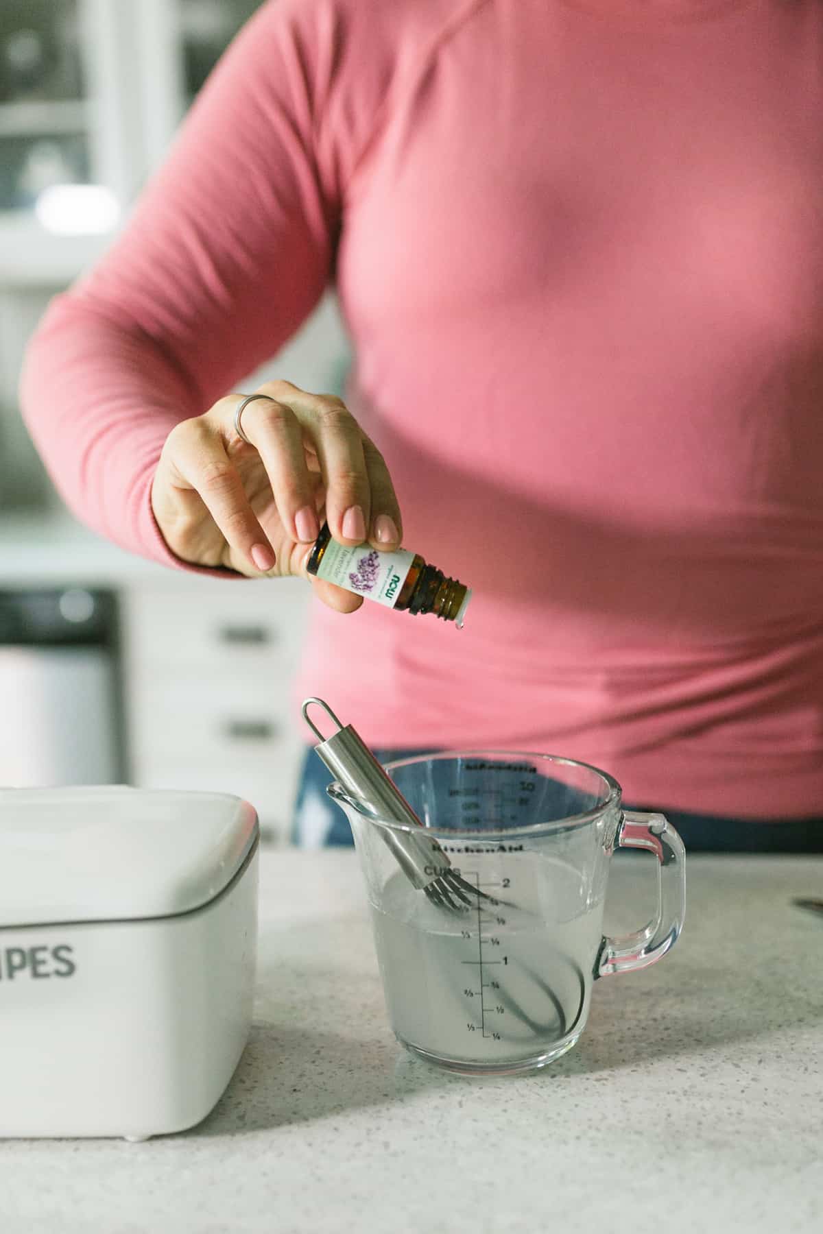 Woman in a pink shirt pouring essential oils into a glass measuring cup that has a whisk sitting in it.