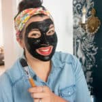 Woman with an activated charcoal face mask on, holding a makeup brush and smiling.