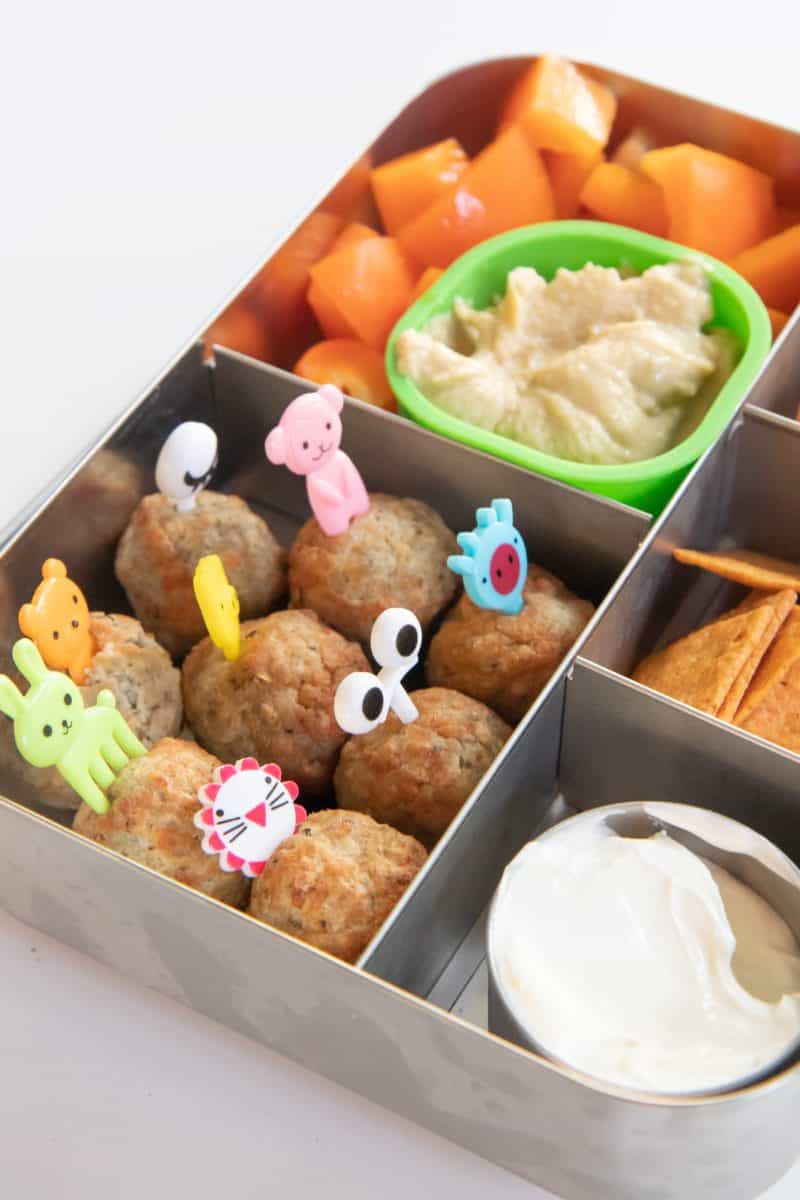 Bento-style lunchbox filled with meatballs, cantaloupe, crackers, and hummus