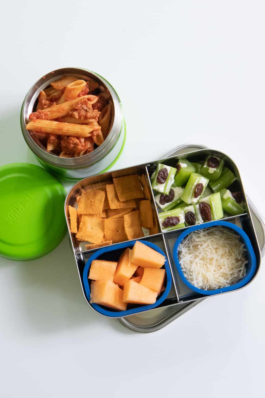 Stainless steel lunch box with cantaloupe, ants on a log, and crackers next to a Thermos of penne in meat sauce.
