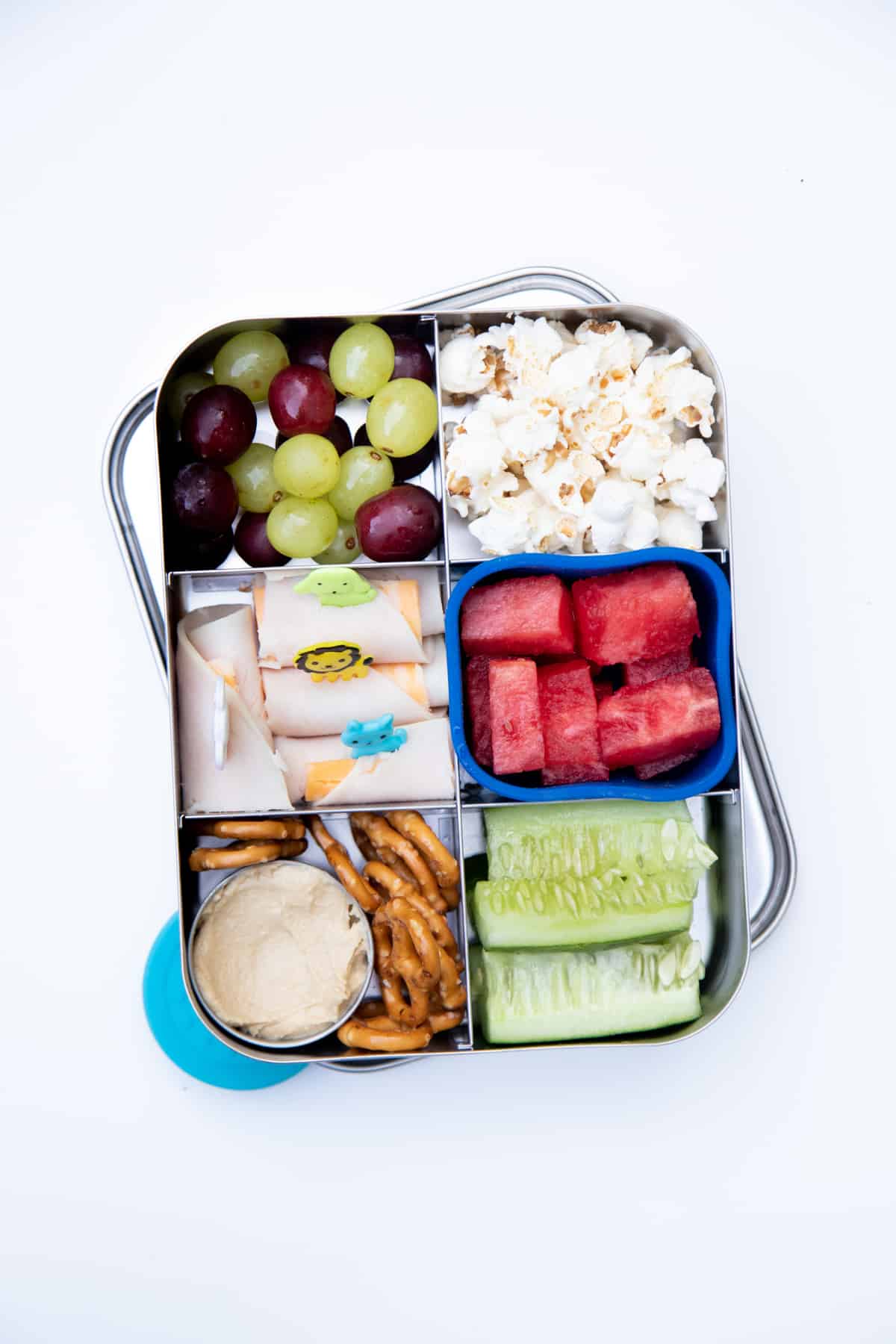 Stainless steel bento-style lunchbox filled with a snack lunch: pretzels and dip, grapes, watermelon, popcorn, cucumber spears, and deli meat roll-ups