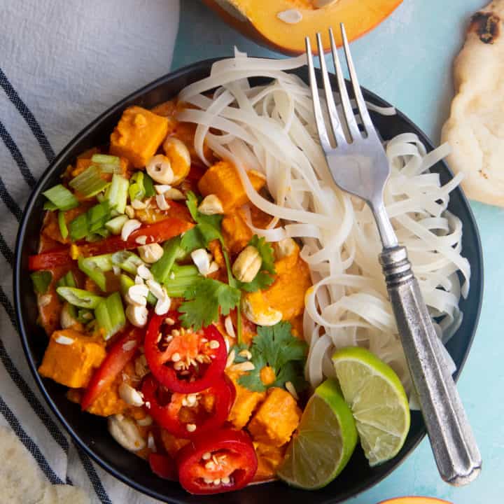 Vegan one pot pumpkin curry served over a bed of rice noodles, in a black dish on a light blue table.