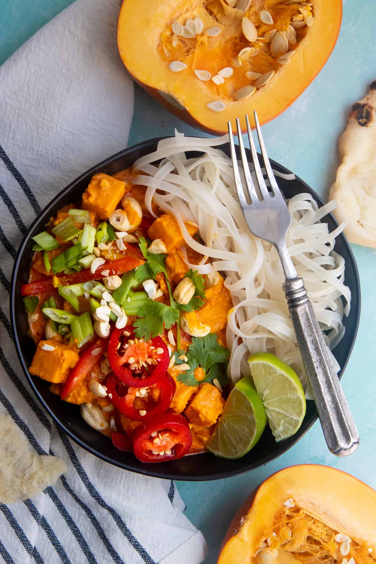 Vegan one pot pumpkin curry served over a bed of rice noodles, in a black dish on a light blue table.
