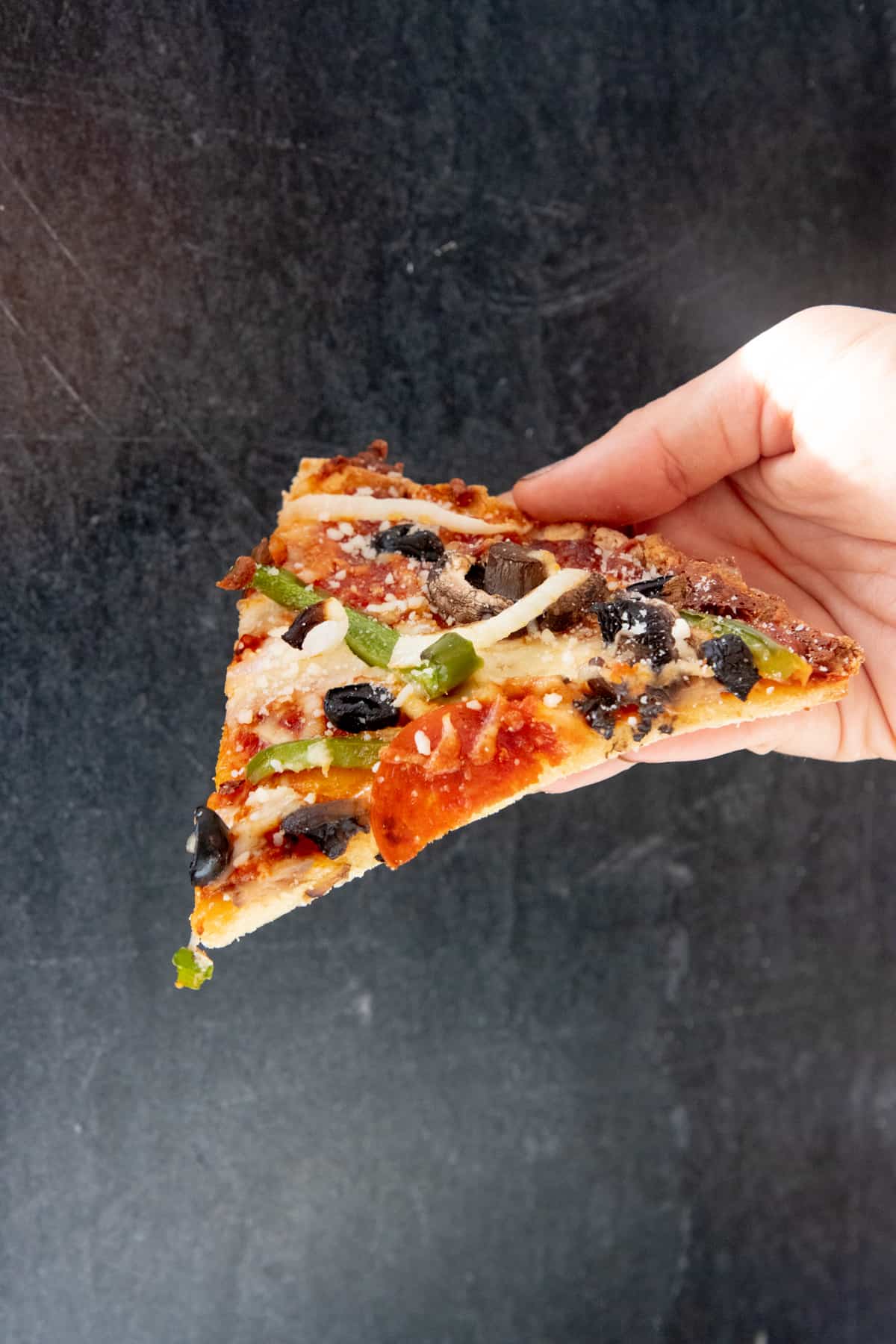 A hand holds a slice of paleo vegetable pizza, in front of a black background.