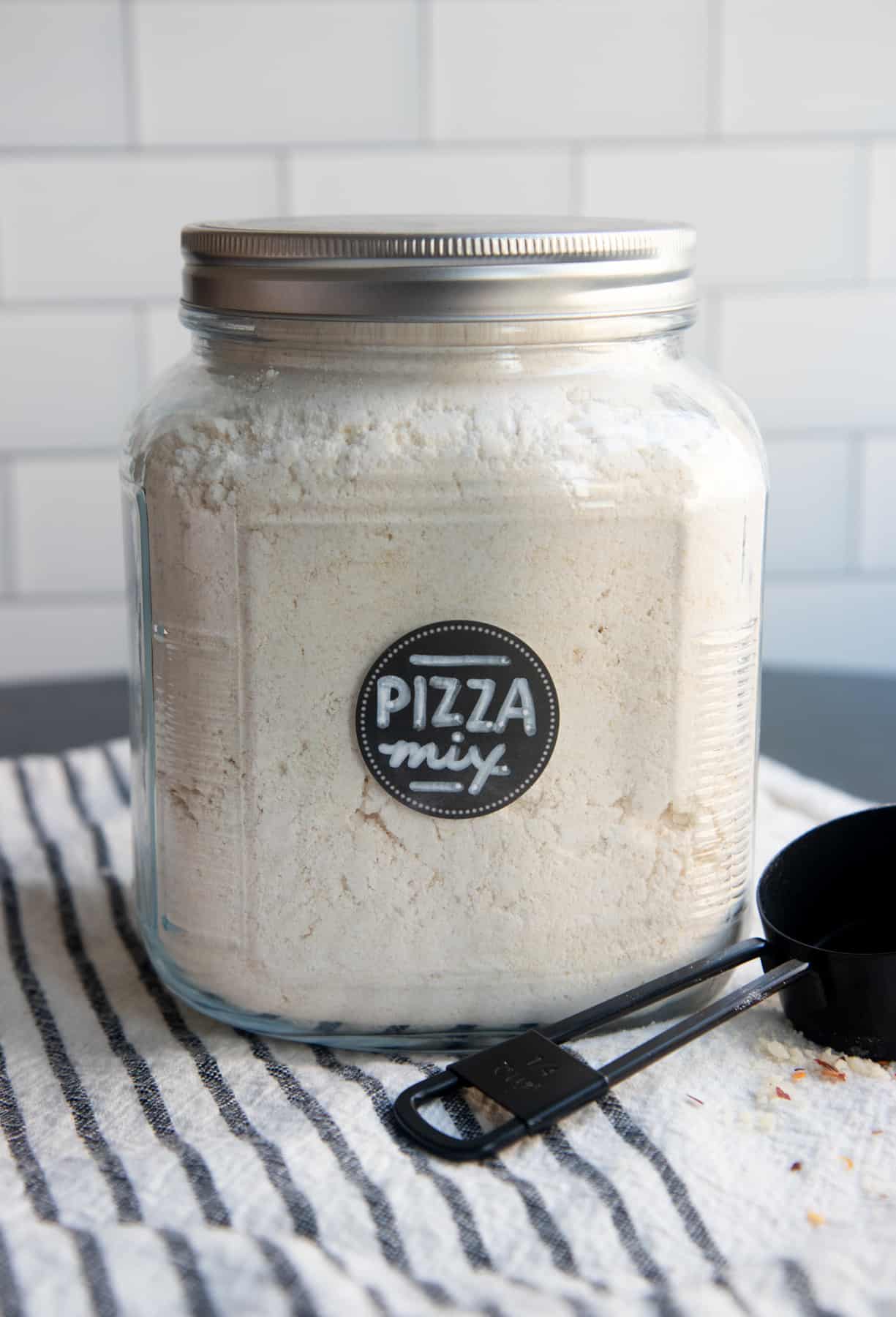 A large open jar of almond flour pizza crust mix sits next to a measuring cup.
