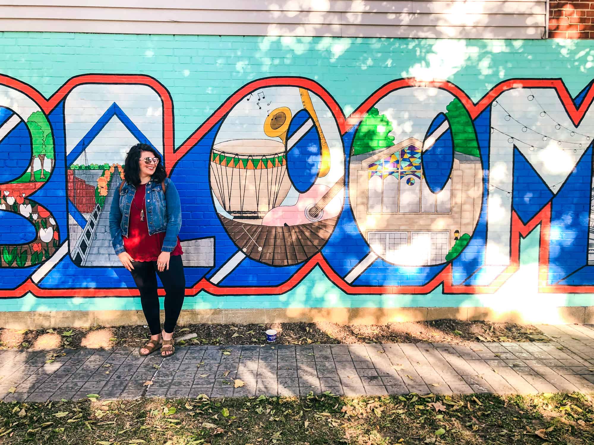 Brunette woman in leggings and a denim jacket stands in front of a painted mural that says "Bloomington," though only the "Bloom" is visible.