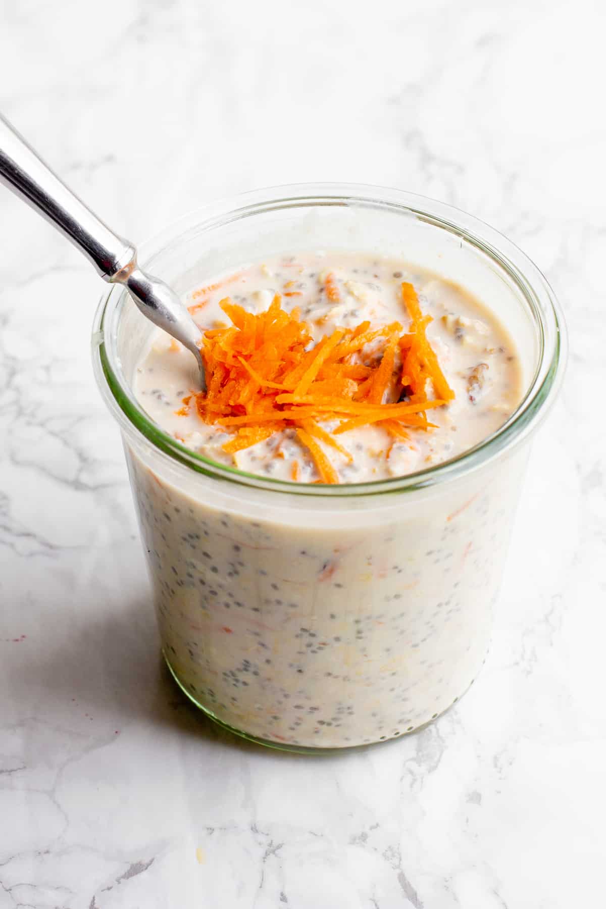 A glass jar filled with carrot cake overnight oats sits on a marble countertop. The oats are garnished with shredded carrots, and a spoon dips into the jar.