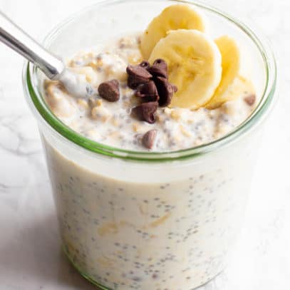 A glass jar filled with chocolate chip banana overnight oats sits on a marble countertop. The oats are garnished with banana slices and chocolate chips, and a spoon dips into the jar.