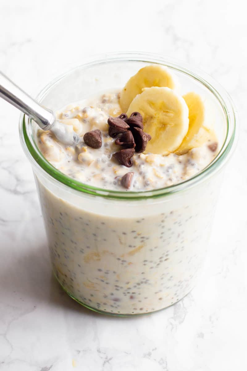 A glass jar filled with chocolate chip banana overnight oats sits on a marble countertop. The oats are garnished with banana slices and chocolate chips, and a spoon dips into the jar.