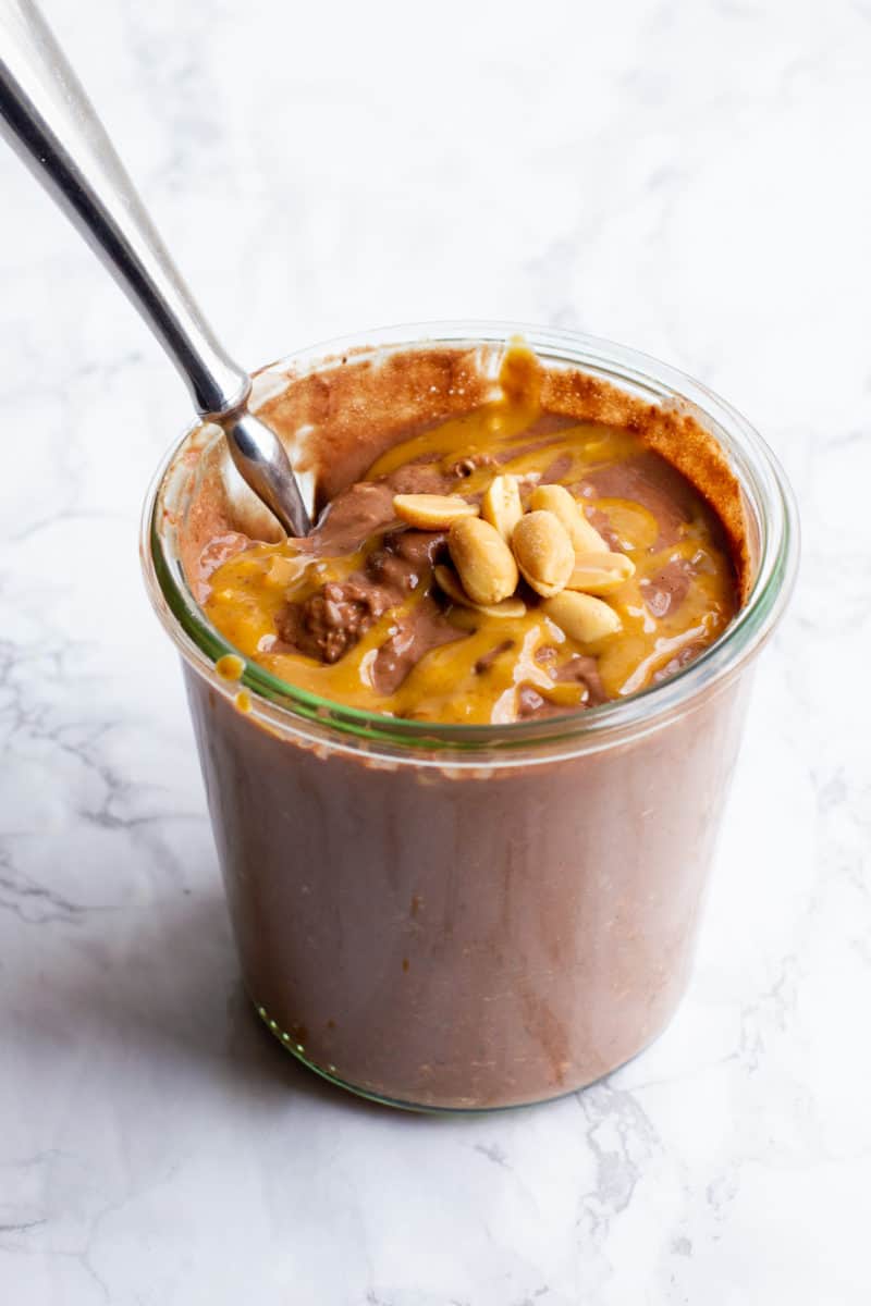 Peanut butter chocolate overnight oats in a glass jar. There are peanuts garnishing the top, and a spoon dips into the jar.