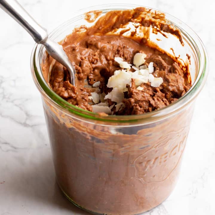 A glass jar filled with coconut chocolate overnight oats sits on a marble countertop. The oats are garnished with a coconut flakes, and a spoon dips into the jar.