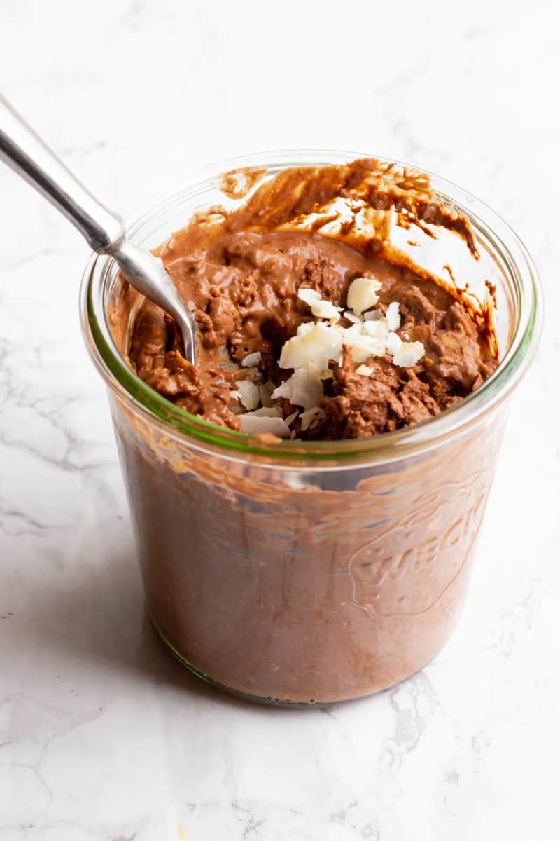 A glass jar filled with coconut chocolate overnight oats sits on a marble countertop. The oats are garnished with a coconut flakes, and a spoon dips into the jar.