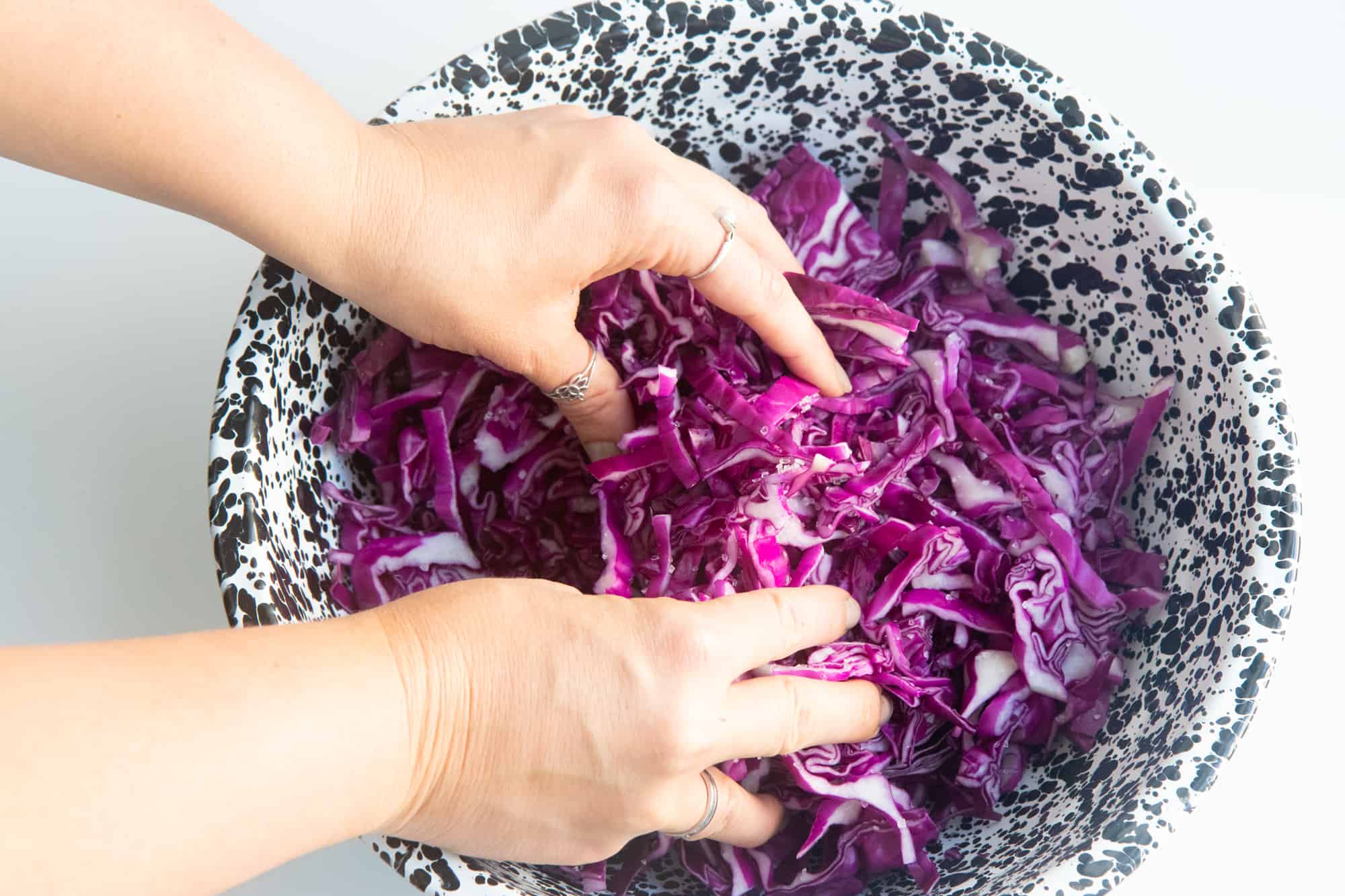 Two hands massage sliced red cabbage in a speckled bowl.