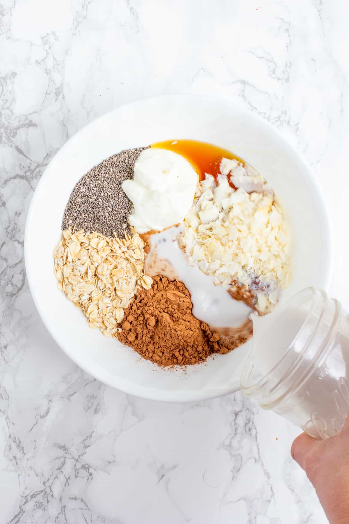 A hand pours coconut milk from a glass jar into a white bowl. The white bowl is filled with other ingredients, including yogurt, cocoa powder, coconut, maple syrup, chia seeds, and rolled oats.