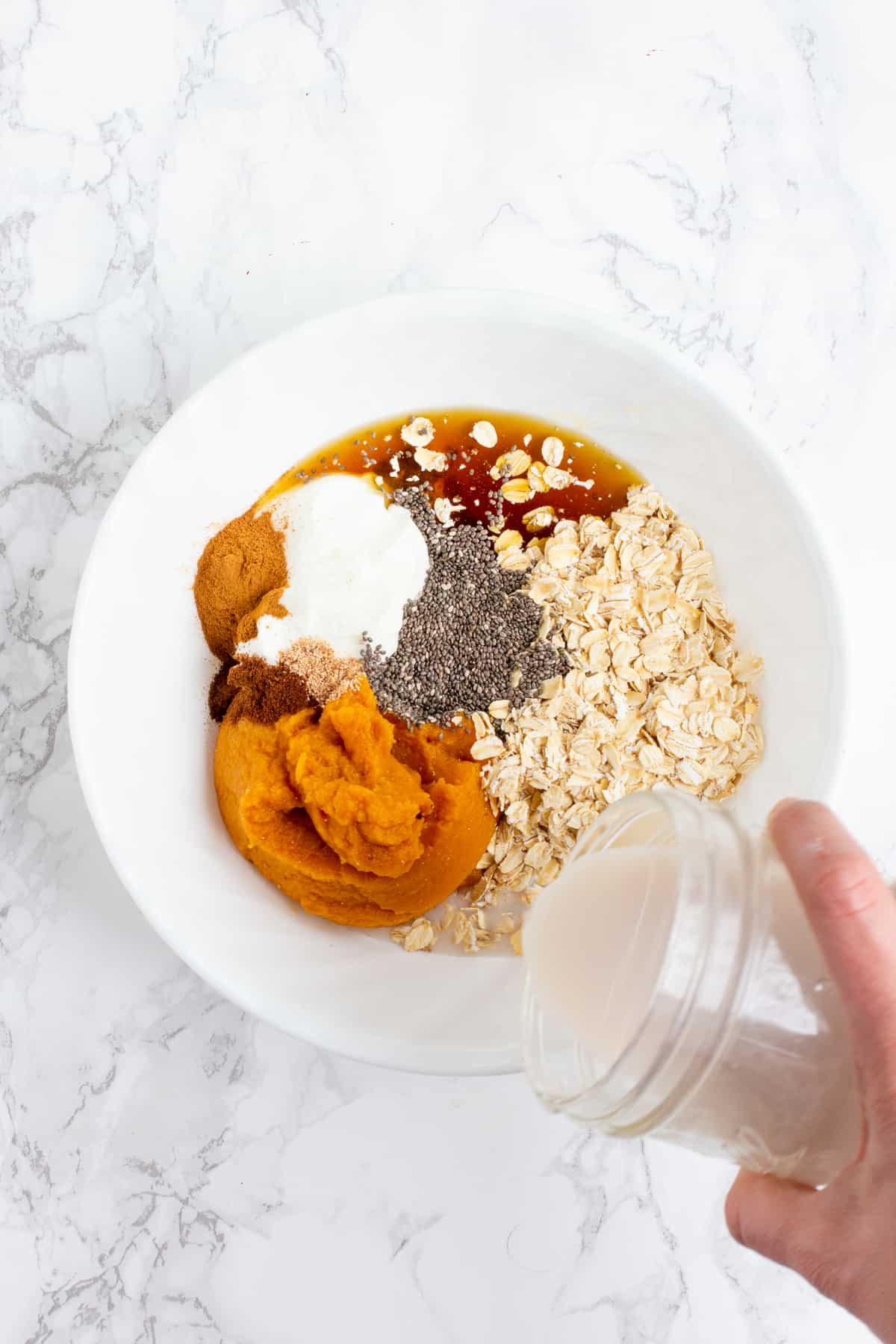 A hand pours milk from a glass jar into a white bowl. The white bowl is filled with other ingredients, including yogurt, pumpkin puree, maple syrup, chia seeds, and rolled oats.