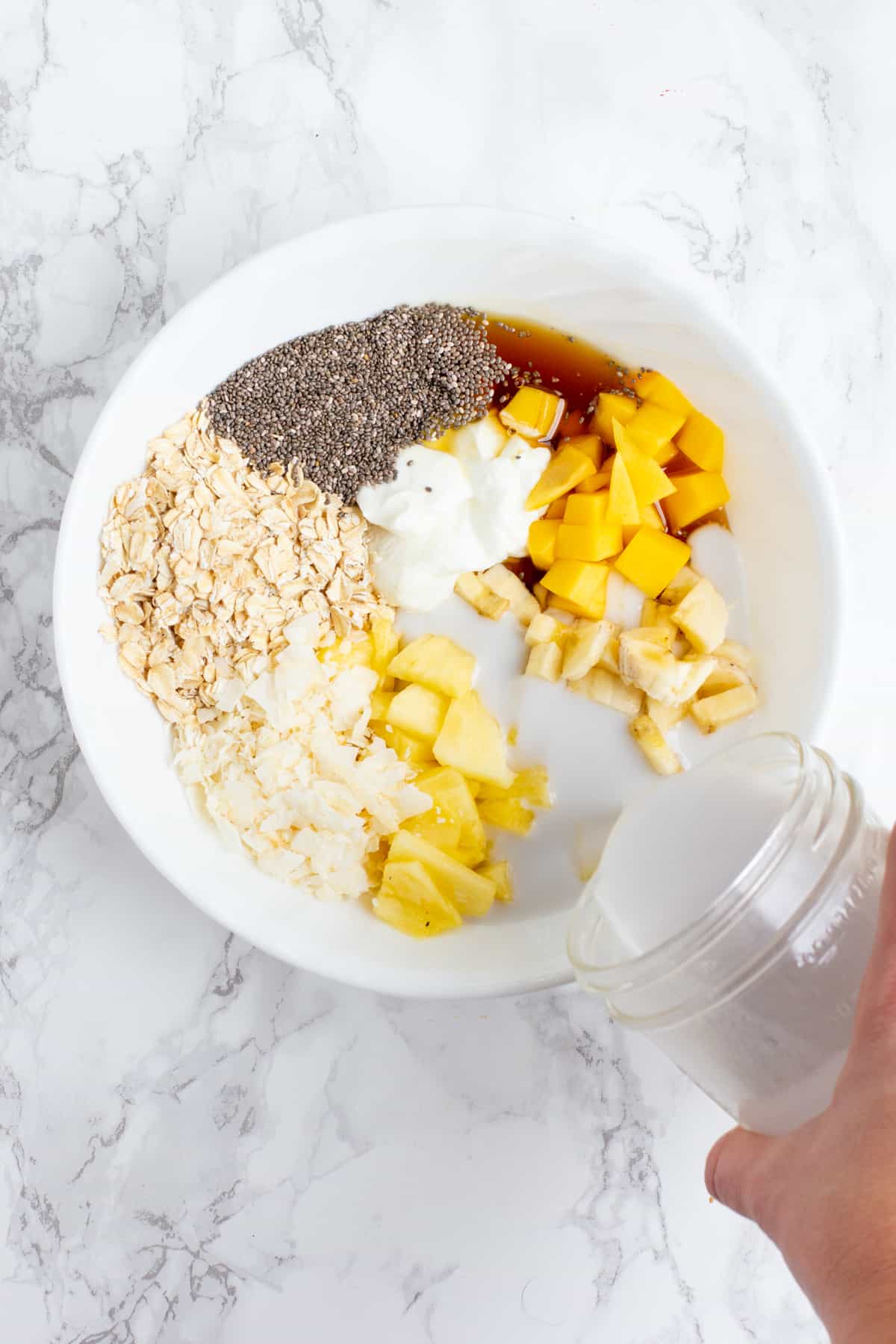 A hand pours milk from a glass jar into a white bowl. The white bowl is filled with other ingredients, including yogurt, chopped pineapple, fresh mango, chia seeds, and rolled oats.