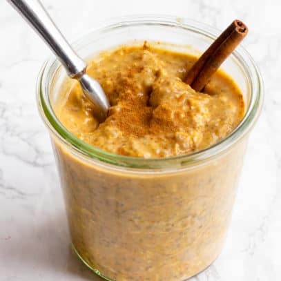 A glass jar filled with pumpkin spice overnight oats sits on a marble countertop. The oats are garnished with a cinnamon stick, and a spoon dips into the jar.