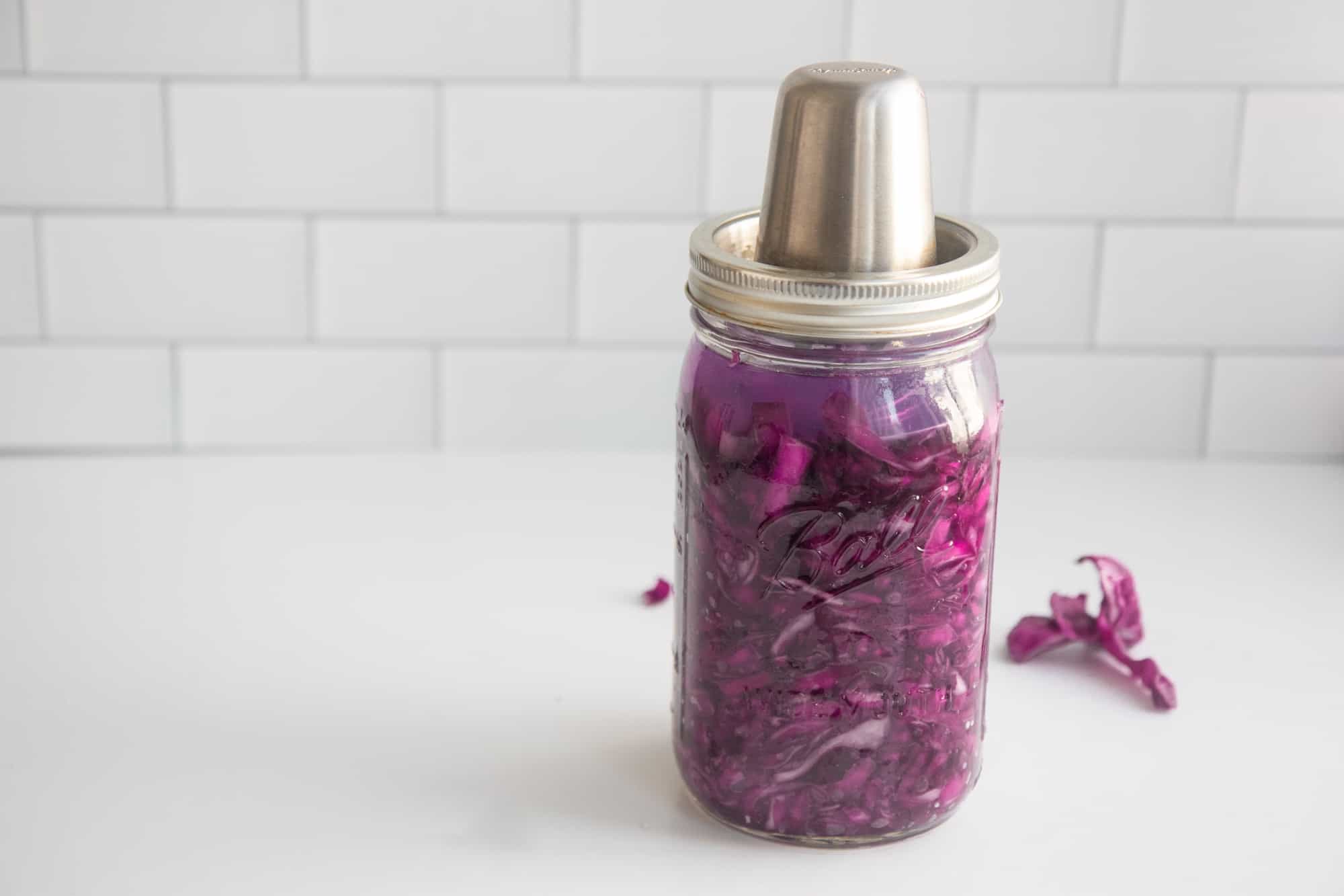 Homemade sauerkraut sits on a counter in a glass jar with a fermentation lid. Red cabbage slices are on the counter behind the jar.
